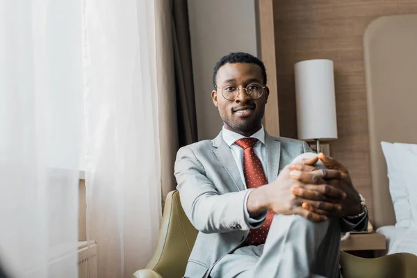 Усміхнений афроамериканський бізнесмен у сірому костюмі сидить у кріслі в готельному номері — Stock Photo