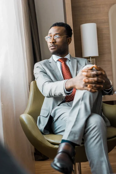 Nachdenklicher afrikanisch-amerikanischer Geschäftsmann im grauen Anzug sitzt im Sessel im Hotelzimmer — Stockfoto