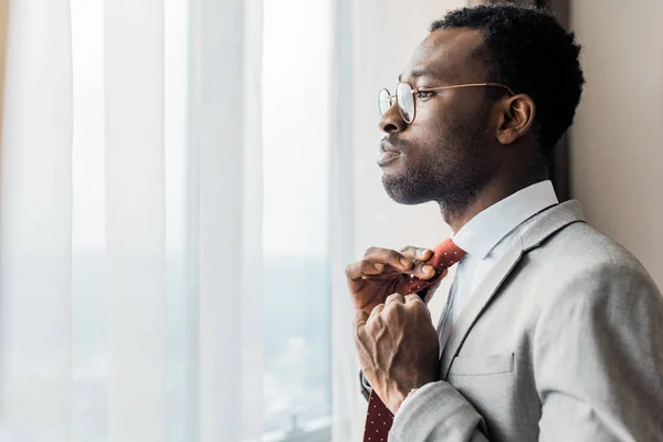 Retrato de perfil do empresário afro-americano ajustando gravata vermelha e olhando para a janela — Fotografia de Stock