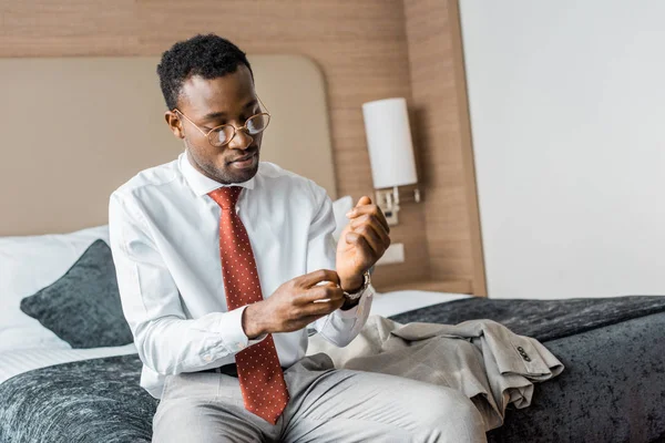 Jovem empresário afro-americano vestindo relógio de pulso enquanto sentado na cama no quarto de hotel — Fotografia de Stock