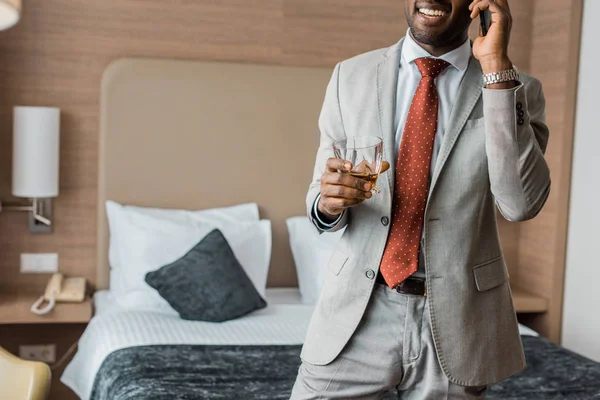 Vista cortada de empresário americano africano alegre com copo de uísque falando no smartphone no hotel — Fotografia de Stock