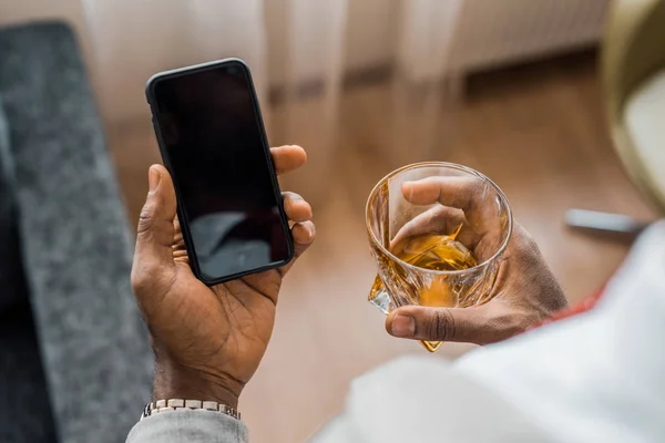 Частичный вид африканского американца со стаканом виски с помощью смартфона — стоковое фото
