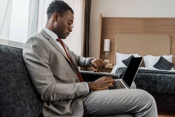 Hombre de negocios afroamericano mirando reloj de pulsera mientras usa el ordenador portátil en la habitación de hotel - foto de stock