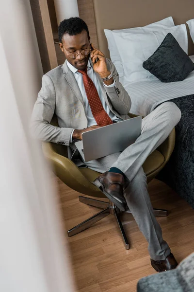 Bonito homem de negócios afro-americano falando no smartphone e usando laptop no quarto de hotel — Fotografia de Stock