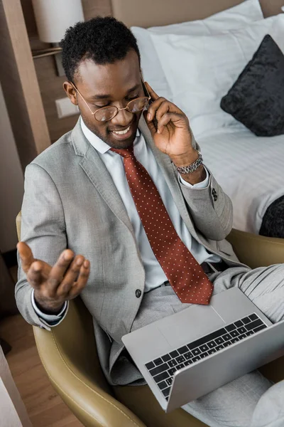 Alegre hombre de negocios afroamericano hablando en el teléfono inteligente y mirando el ordenador portátil en la habitación de hotel - foto de stock