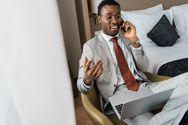 Bem sucedido bonito empresário afro-americano com laptop falando no smartphone no quarto de hotel — Fotografia de Stock