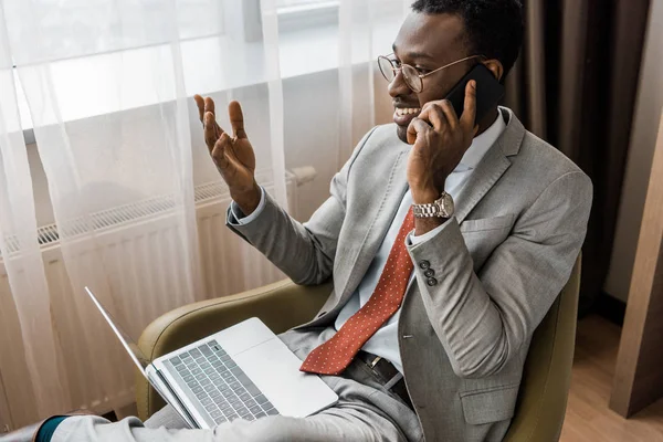Uomo d'affari afroamericano sorridente gesticolando, parlando su smartphone e utilizzando il computer portatile in camera d'albergo — Foto stock