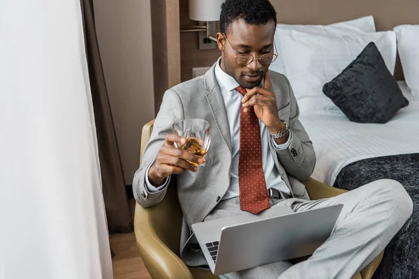 Pensativo hombre de negocios afroamericano con vaso de coñac mirando portátil en habitación de hotel - foto de stock