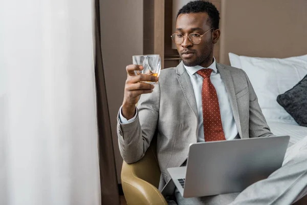 Handsome african american businessman with laptop looking at glass of cognac in hotel — Stock Photo