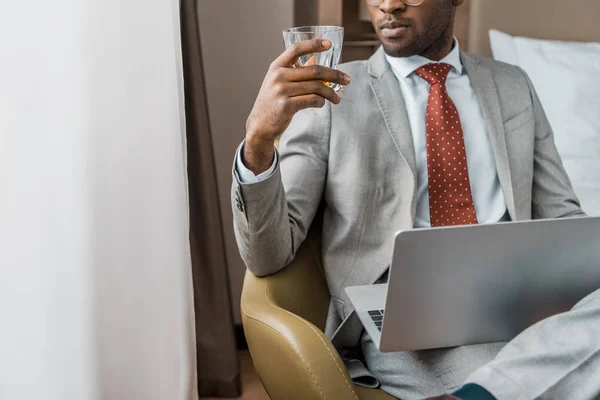 Vista recortada de hombre de negocios afroamericano con portátil mirando el vaso de bebida alcohólica en el hotel - foto de stock