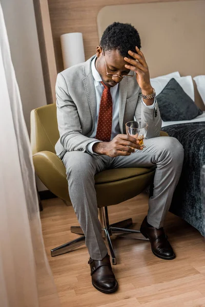 Frustrated african american man holding glass of cognac in hotel room — Stock Photo