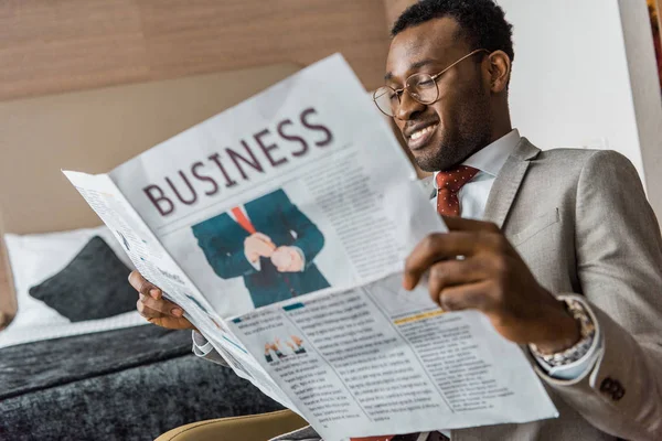 Sonriente hombre de negocios afroamericano en traje leyendo periódico de negocios en habitación de hotel - foto de stock