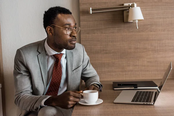 Hombre de negocios afroamericano sosteniendo taza de café en la mesa con el ordenador portátil - foto de stock