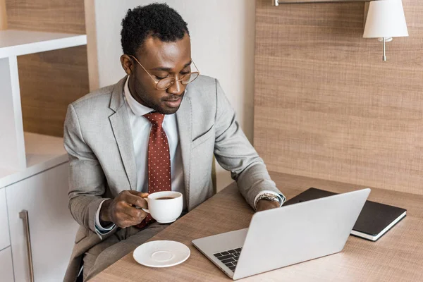 Homme d'affaires afro-américain en costume tenant tasse de café tout en travaillant sur ordinateur portable dans la chambre d'hôtel — Photo de stock
