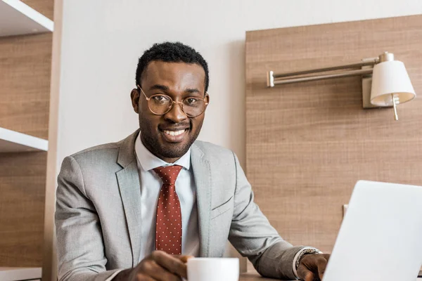 Sonriente hombre de negocios afroamericano con portátil en la pausa de café en la habitación de hotel - foto de stock