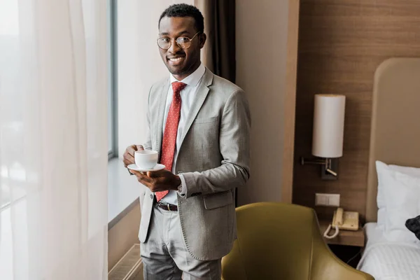 Alegre afroamericano hombre de negocios sosteniendo taza de café en habitación de hotel - foto de stock