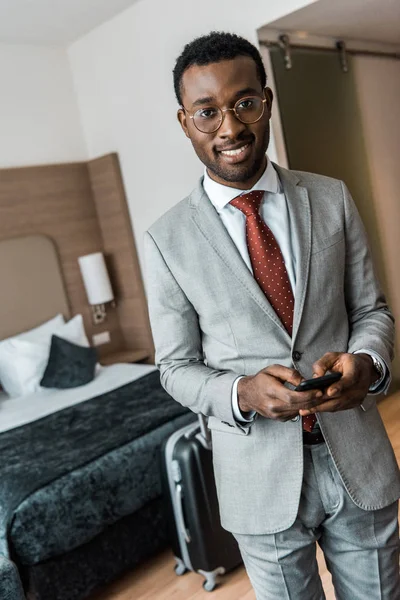 Uomo d'affari afroamericano sorridente utilizzando smartphone in camera d'albergo — Foto stock