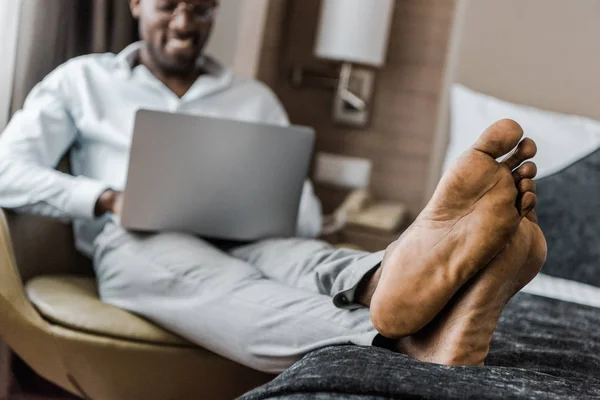 Enfoque selectivo del hombre afroamericano descalzo usando el ordenador portátil en la habitación de hotel - foto de stock