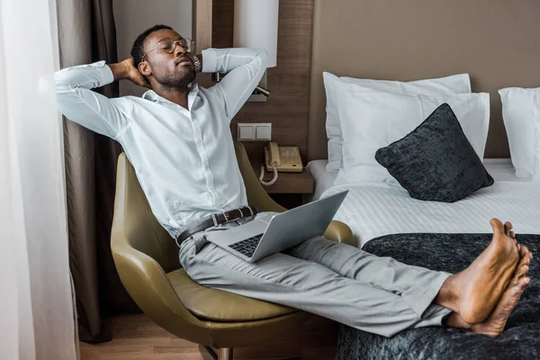 Homme afro-américain pieds nus avec les yeux fermés relaxant dans un fauteuil avec ordinateur portable dans la chambre d'hôtel — Photo de stock