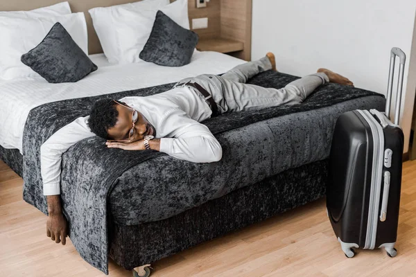Hombre afroamericano cansado en ropa formal durmiendo en la cama en la habitación de hotel con maleta - foto de stock