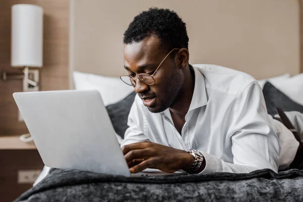 Bel homme d'affaires afro-américain concentré travaillant avec ordinateur portable sur le lit de l'hôtel — Photo de stock