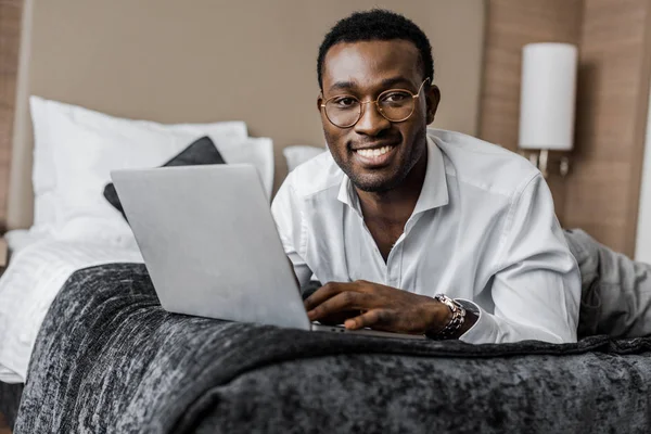 Heureux homme d'affaires afro-américain en lunettes à l'aide d'un ordinateur portable sur le lit de l'hôtel — Photo de stock