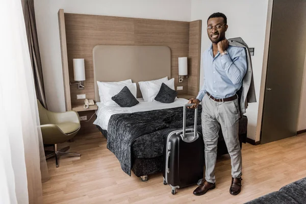 Smiling african american businessman with suitcase in hotel room with bed — Stock Photo