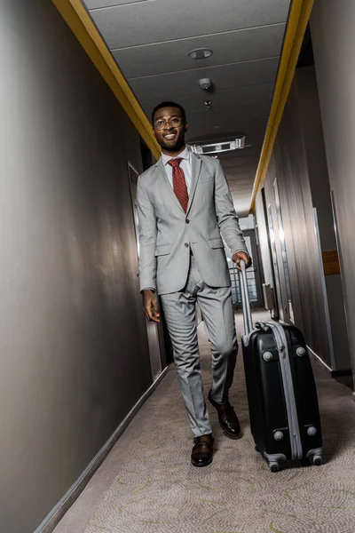 Sonriente hombre de negocios afroamericano en traje caminando con bolsa de viaje en el pasillo del hotel - foto de stock