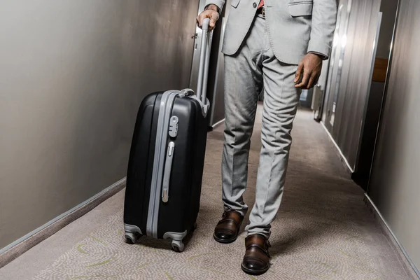 Vista recortada de hombre de negocios en traje gris con maleta caminando en el pasillo del hotel - foto de stock