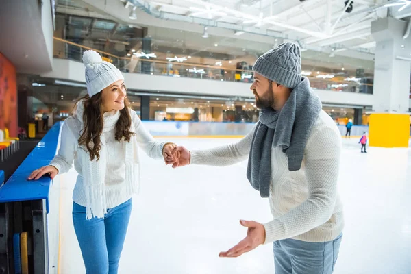 Coppia sorridente in cappelli e maglioni che si tiene per mano mentre pattina sulla pista di pattinaggio — Foto stock