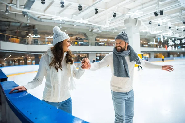 Smiling couple in hats and sweaters holding hands while skating on ice rink — Stock Photo