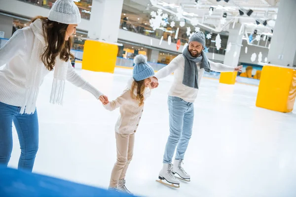 Familia feliz en sombreros y suéteres tomados de la mano mientras patinan juntos en pista de hielo - foto de stock