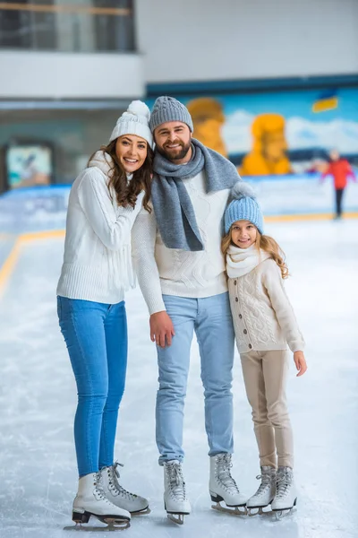Pais sorridentes e filha em camisolas olhando para a câmera na pista de patinação — Fotografia de Stock