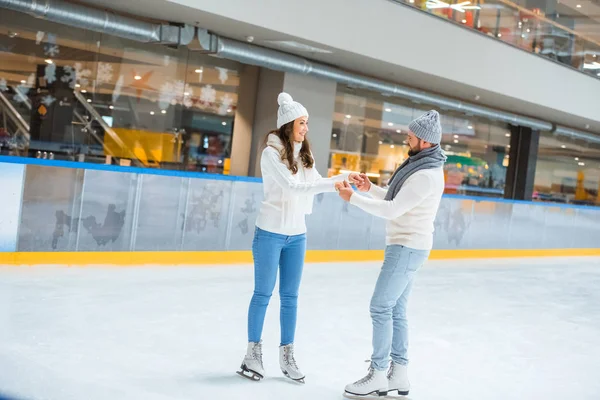 Coppia sorridente in cappelli e maglioni che si tiene per mano mentre pattina sulla pista di pattinaggio — Foto stock