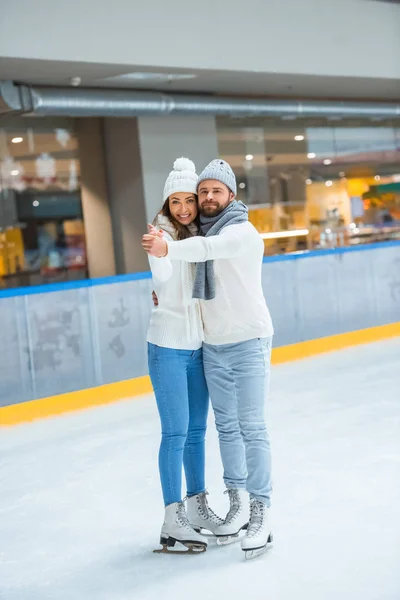 Sorrindo casal em chapéus de malha e camisolas abraçando e de mãos dadas na pista de patinação — Fotografia de Stock