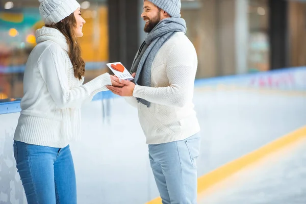 Vista lateral do homem cumprimentando namorada com cartão postal na pista de patinação, st Valentine conceito dia — Fotografia de Stock