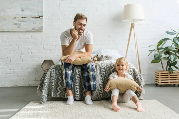 Happy father and daughter holding pillows and smiling at camera in bedroom — Stock Photo