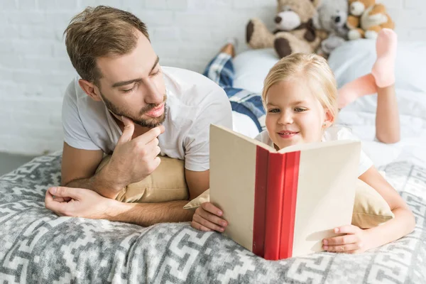 Батько дивиться на милу маленьку доньку, читаючи книгу на ліжку — стокове фото