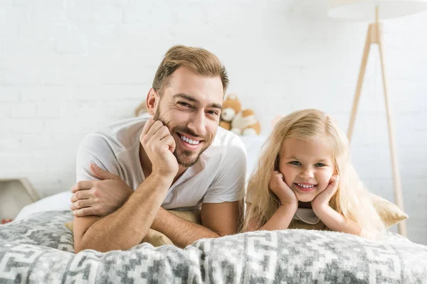 Счастливый отец и милая маленькая дочь лежат вместе на кровати и улыбаются в камеру — стоковое фото