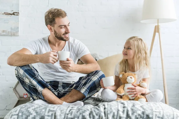 Happy father and daughter holding tea cups and smiling each other on bed — Stock Photo