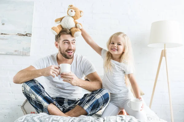 Милая маленькая дочь играет с плюшевым мишкой и улыбается в камеру, пока счастливый отец пьет чай на кровати — стоковое фото