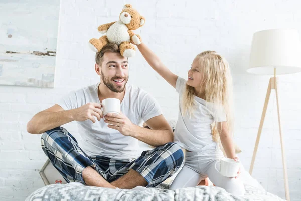 Smiling little daughter playing with teddy bear while happy father drinking tea in bedroom — Stock Photo