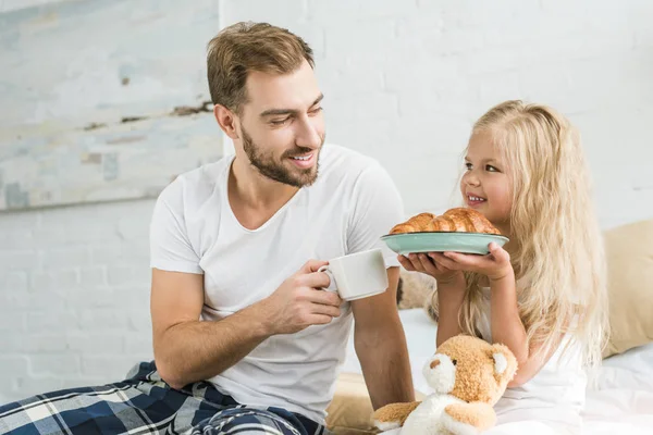 Pai feliz em pijama segurando xícara de chá e olhando para a pequena filha com croissants saborosos no prato — Fotografia de Stock