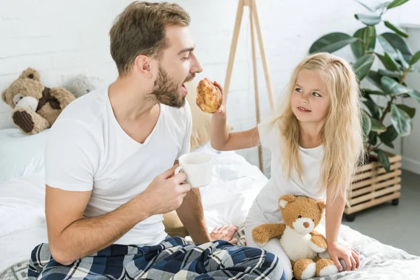 Niedliche kleine Tochter füttert Vater mit Croissant im Bett — Stockfoto