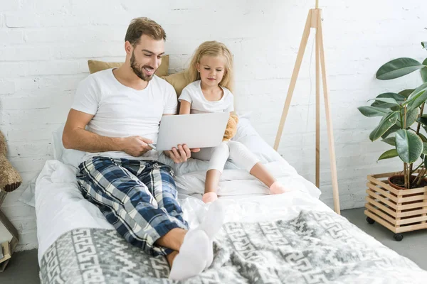 Vista de ángulo alto de padre feliz y linda hija pequeña usando el ordenador portátil juntos en la cama - foto de stock