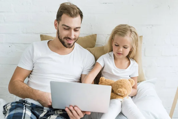 Glücklicher Vater und süße kleine Tochter mit Teddybär mit Laptop zusammen im Schlafzimmer — Stockfoto