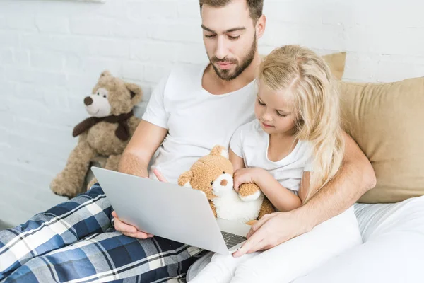 Padre y adorable hijita usando el ordenador portátil juntos en la cama - foto de stock