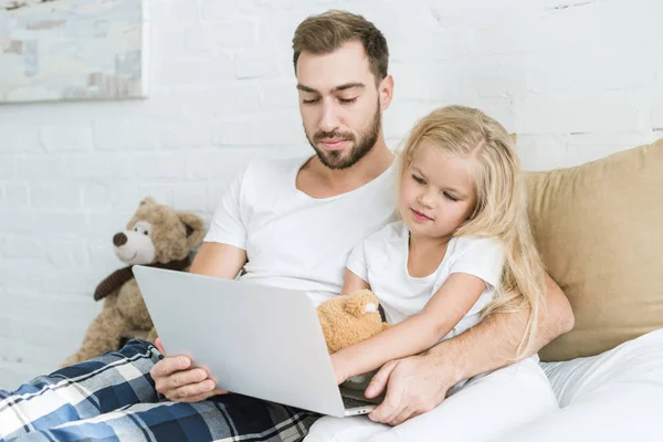 Padre y linda hijita con oso de peluche utilizando el ordenador portátil juntos en el dormitorio - foto de stock