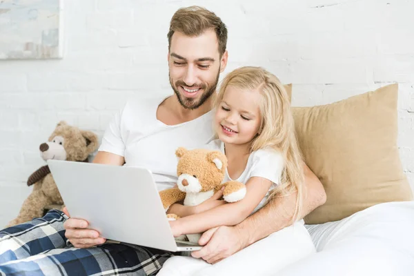 Feliz padre y linda hijita usando el ordenador portátil juntos en el dormitorio - foto de stock