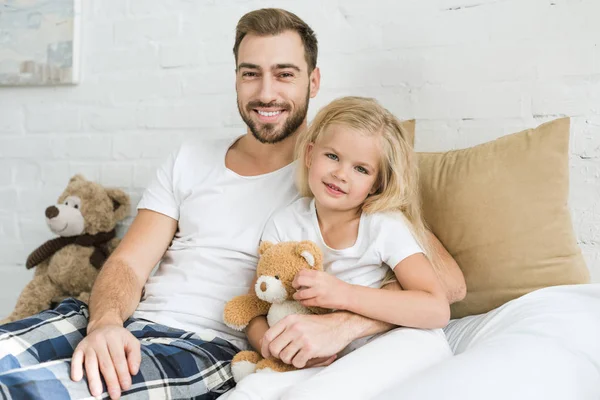 Feliz padre y linda hijita con oso de peluche sentado en la cama y sonriendo a la cámara - foto de stock